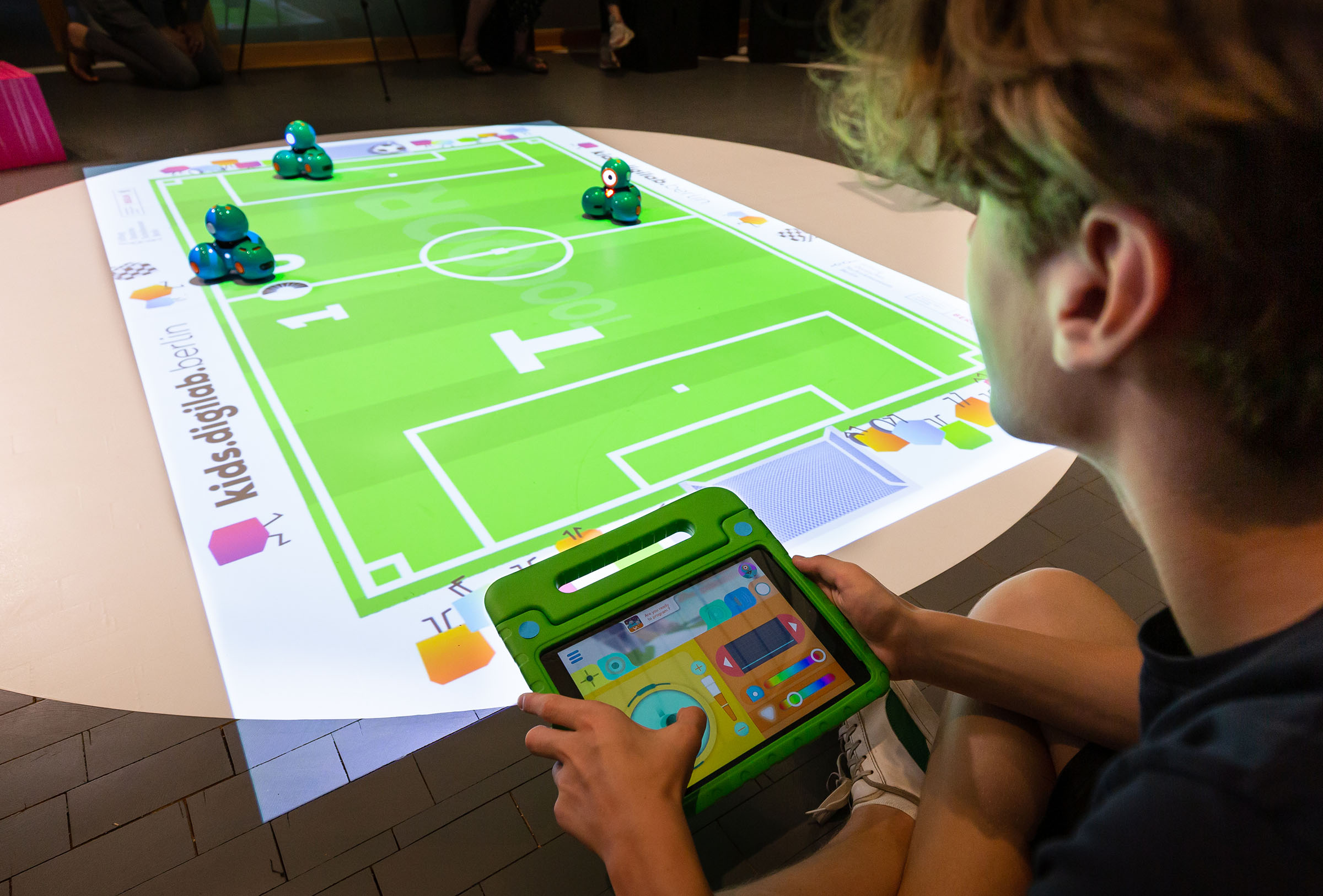 Drei Dash-Roboter spielen auf einem projizierten Fußballfeld Fußball. Ein Kind steuert mit Tablet einen Roboter.