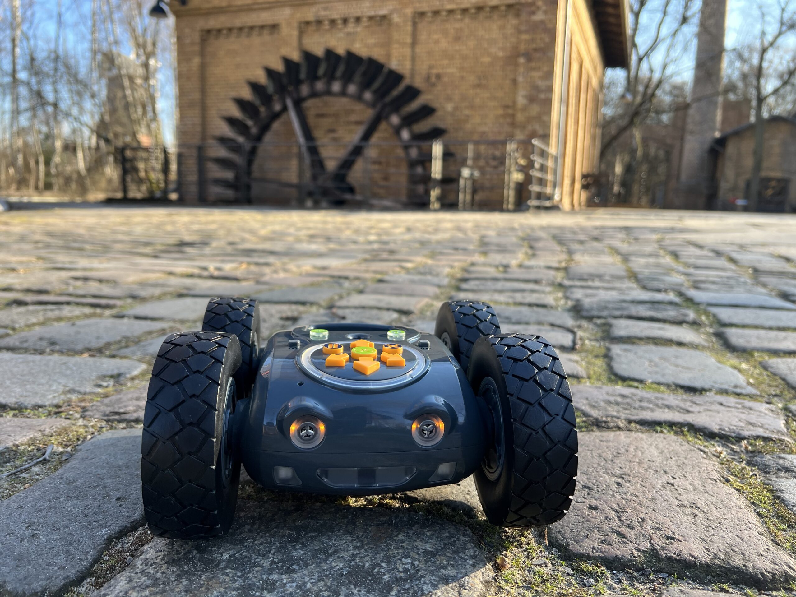Der Rugged Robot steht auf Kopfsteinpflaster im Hintergrund ist ein Gebäude und ein Mühlenrad zu sehen.